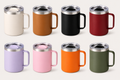 insulated travel mugs with handles and stainless steel lids, shown in assorted colors on a light neutral background. The mugs include cream, tan, black, red, lavender, pink, orange, and olive green.